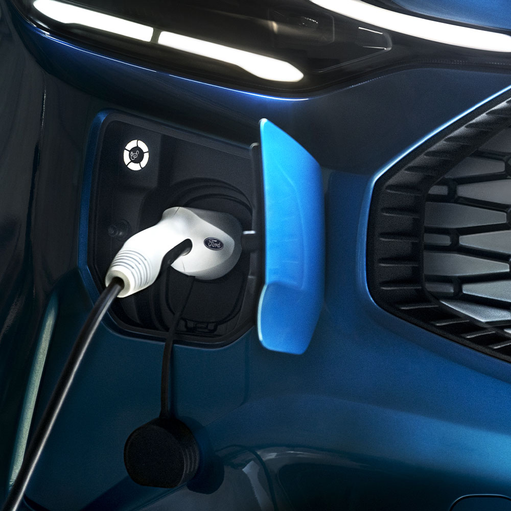 Un puerto de carga para coche eléctrico con un cargador de coche eléctrico conectado en primer plano que contrasta con un fondo en el que se ve un coche Ford azul.