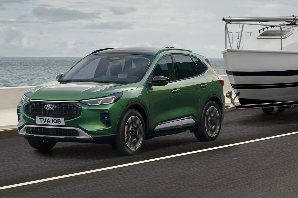 Ford Kuga verde remolcando un pequeño barco por una carretera de la costa.