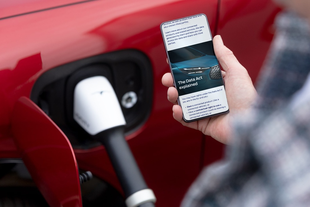 Imagen de una persona revisando su móvil con un vehículo eléctrico rojo cargando en segundo plano.