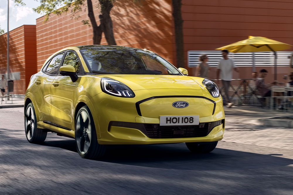 Vista frontal de un Ford Puma Gen-E® eléctrico amarillo circulando por una carretera urbana.