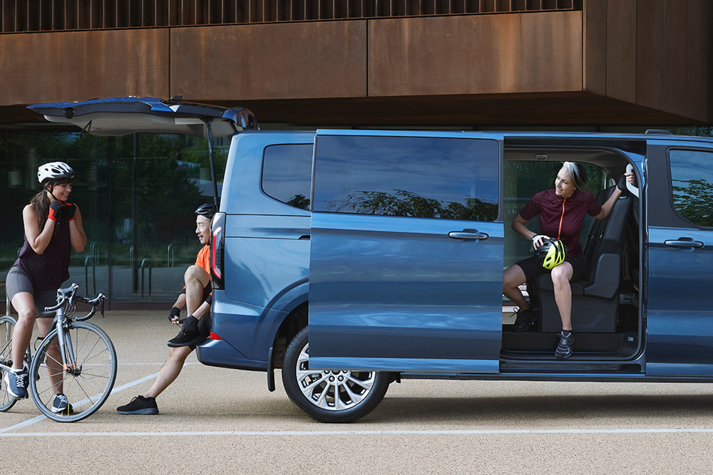 Ford Tourneo Custom azul aparcada con las puertas laterales abiertas y un grupo de personas con bicicletas cerca. 