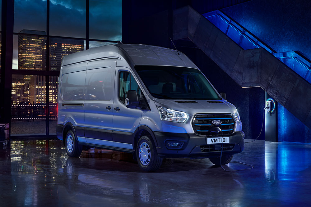 Furgoneta eléctrica Ford E-Transit en color plateado aparcada en el interior de un recinto desde el que se ve la línea del horizonte de una ciudad a través de grandes ventanales. 