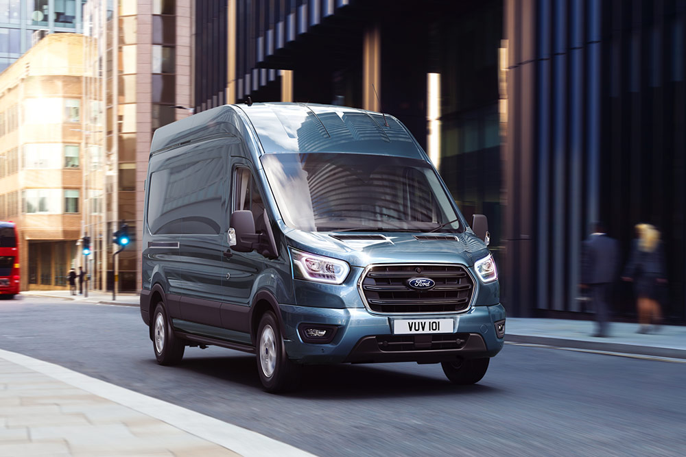 Una furgoneta Ford Transit azul grisácea circulando por una calle de la ciudad.