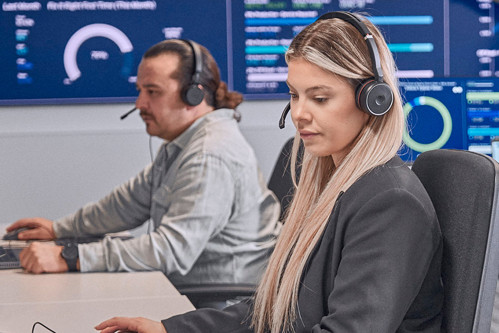 Dos personas con auriculares concentradas en pantallas de ordenador en una sala de control moderna, con datos.