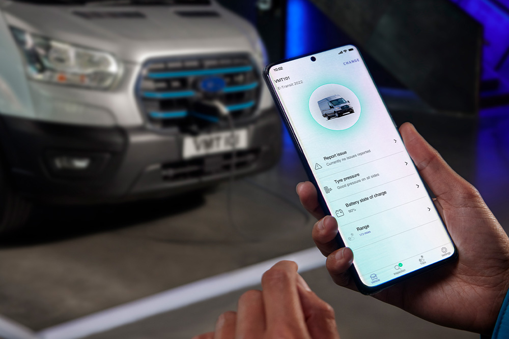 Una persona sosteniendo un smartphone que muestra una interfaz de aplicación, con una furgoneta Ford Pro Transit plateada difuminada al fondo.