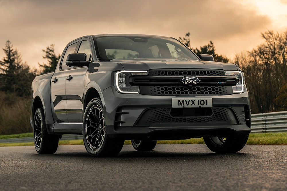 Ford Pro Ranger Híbrido Enchufable gris aparcado en una carretera mojada, con árboles al fondo.