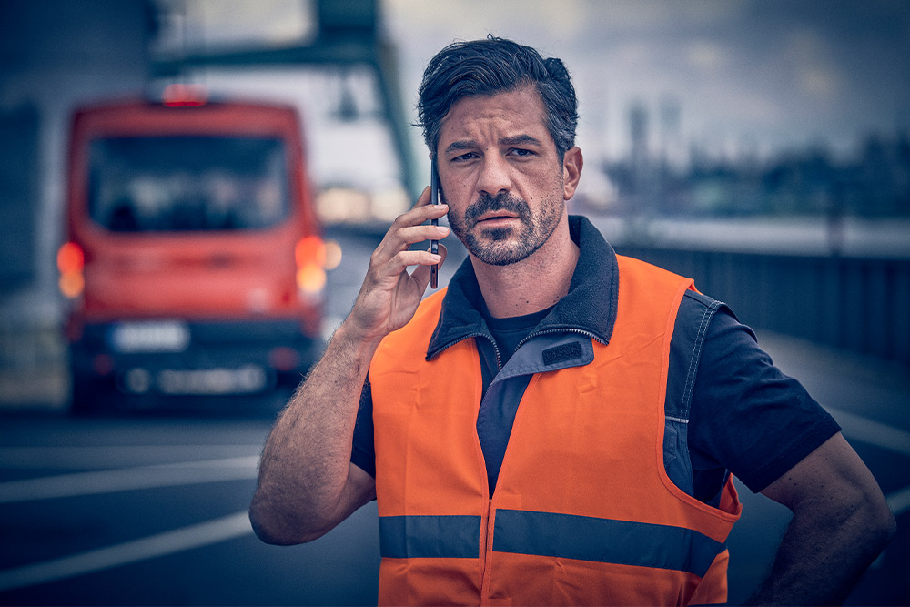 Trabajador con chaleco naranja llamando a asistencia en carretera en un dispositivo móvil.