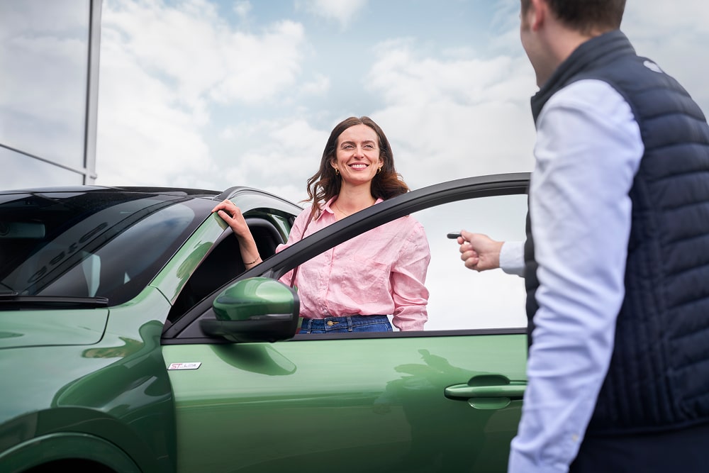Una mujer con una camisa rosa aparece sonriendo junto a un coche verde, con un trabajador del concesionario Ford entregándole las llaves de su coche.