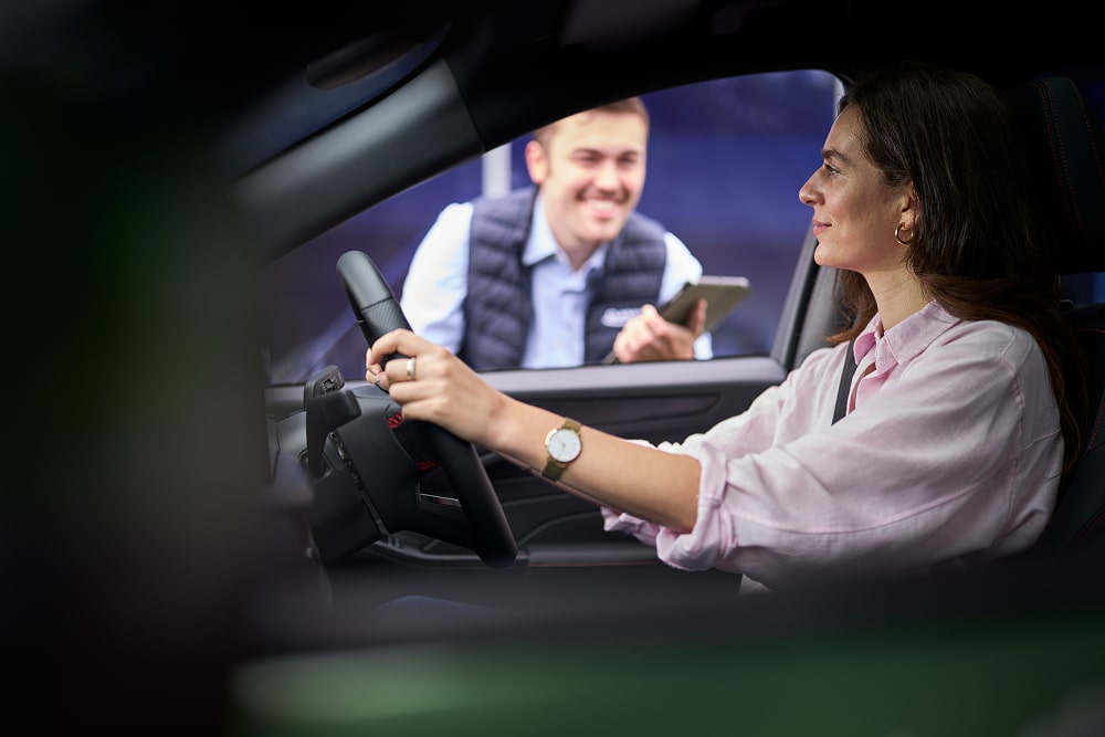 Una mujer en un coche verde aparcado aparece sonriendo por la ventana a un trabajador de un concesionario Ford.