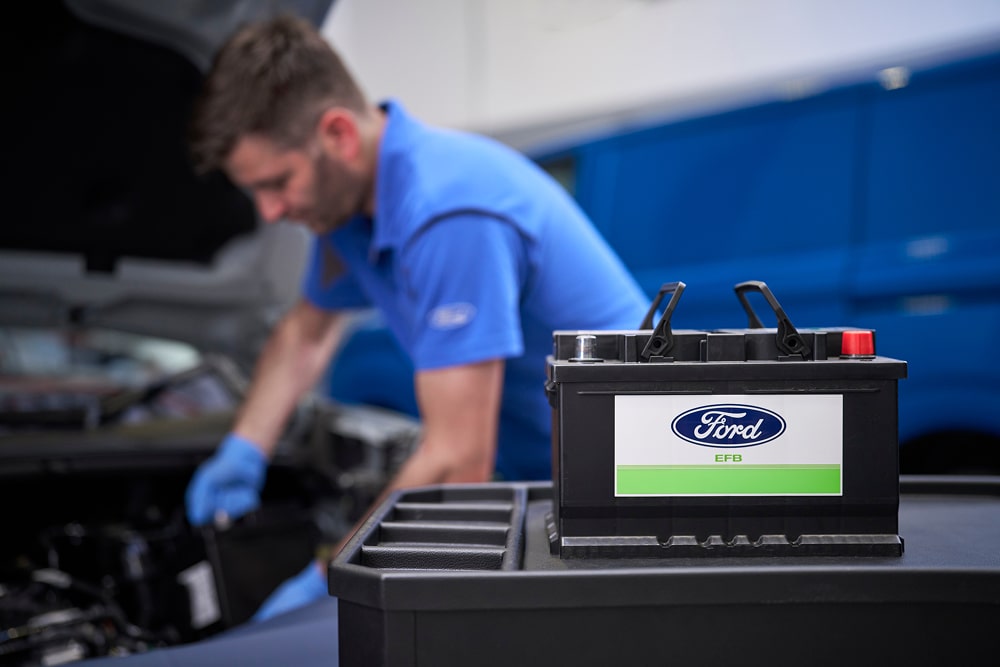 Un mecánico de Ford trabaja en un motor de un coche. Se ve una batería de coche Ford en primer plano.