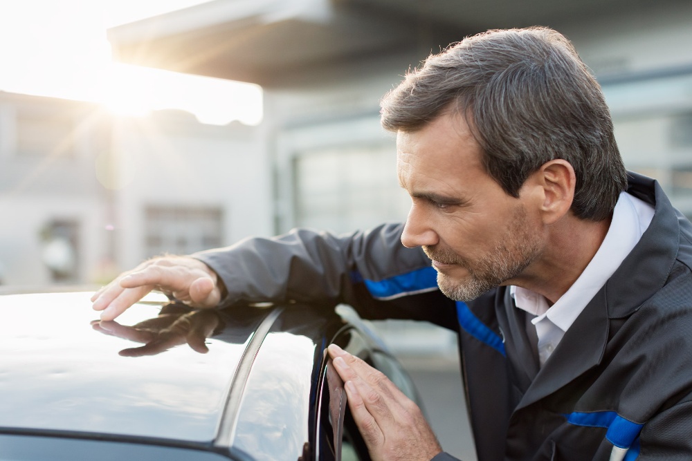 A technician inspects the roof of a car, with sunlight reflecting off the surface.