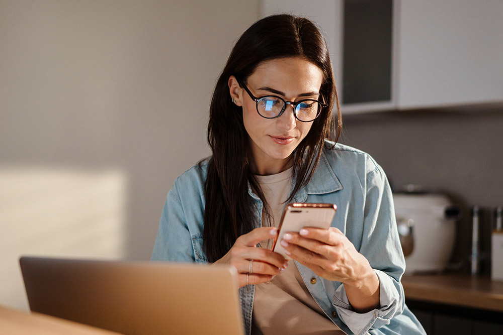 Person wearing glasses looking at a smartphone, with a laptop on a wooden table.